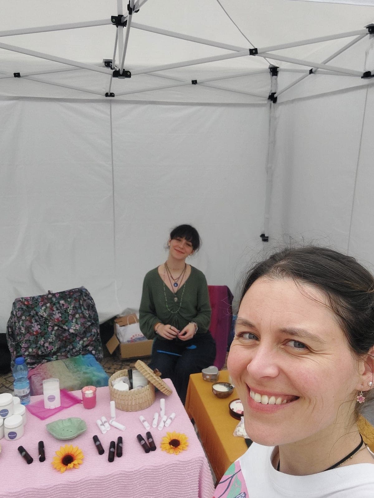 duas mulheres a sorrir dentro de uma tenda branca, com uma mesa rosa de produtos lucrescente e girassóis